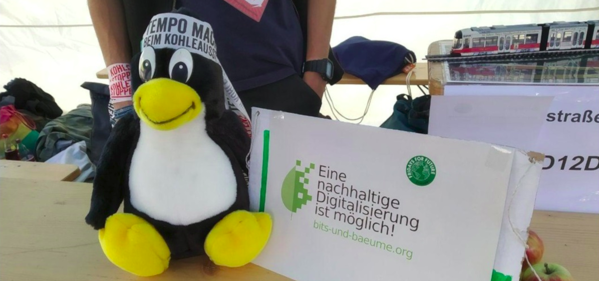 Ein Pinguin-Plüschtier mit einem Stirnband mit verschiedenen Klimaschutz-Sprüchen darauf, um. Daneben ein Schild auf dem: Eine nachhaltige Digitalisierung ist möglich, steht und die Logos von Bits und Bäume und Fridays for Future abgebildet sind. Bits und Bäume Dresden