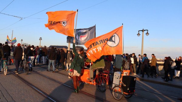 Eine Gruppe Menschen mit Fahnen und Lastenrad inmitten einer Demonstration auf einer Brücke. Die Fahnen sind von der Piratenpartei und Antifa-Fahnen. Brandmauer_600