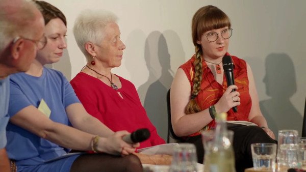 Mehrere Menschen auf einem Podium. Rechts ich, rotbraune Ponyfrisur mit Zopf, große runde Brille, blass, rot-orange Bluse, auf einer Bühne mit Mikrofon in der Hand. Dear_Future