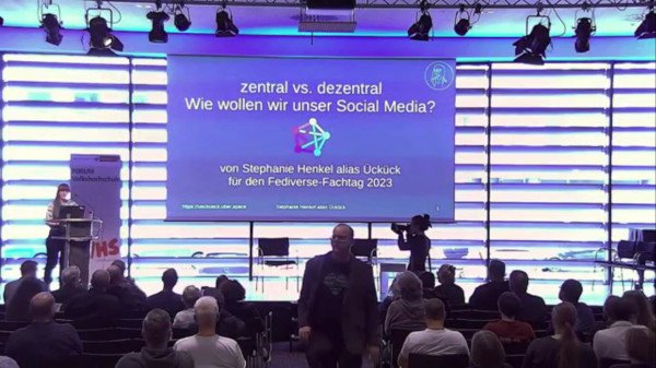 Ein unscharfes Bild mit einer Person am Sprecher*innen-Pult und Publikum. Eine Präsentationsfolie im Hintergrund. Auf dieser grünen Folie ist das Fediverse-Logo in der Mitte angedeutet. Der Titel lautet: zentral vs. dezentral Wie wollen wir uns Social Media? Fediverse-Fachtag