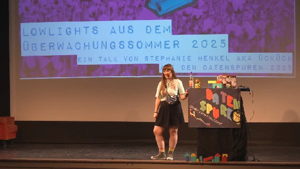 Ich, rotbraune Ponyfrisur, lange Haare, blass, große Brille, auf einer Bühne neben einem Pult. Vorschau_%C3%9Cberwachungssommer